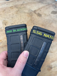 Thumbnail for Two black magazine pouches labeled '300 Blackout' and '5.56 NATO' held by a hand on a wooden surface.